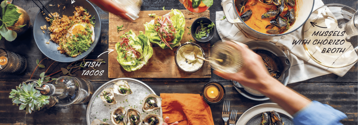 Top-down view of a seafood-based meal spread with hands hands actively cheers-ing